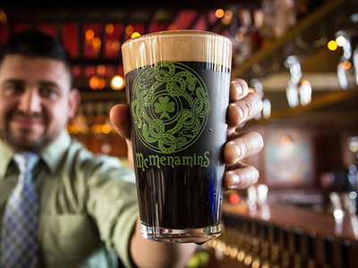 A man in a green shirt and tie holding out a glass of Guinness beer in a McMenamins Pub glass