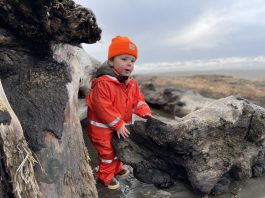 little boy in a orange rain suit and orange beanie stands among driftwood on an Olympic beach.