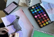 a child's hands are seen painting a goat on paper from a photo a on a phone. a paint set is above the painting and a green paper pad is next to it.