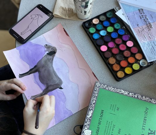 a child's hands are seen painting a goat on paper from a photo a on a phone. a paint set is above the painting and a green paper pad is next to it.
